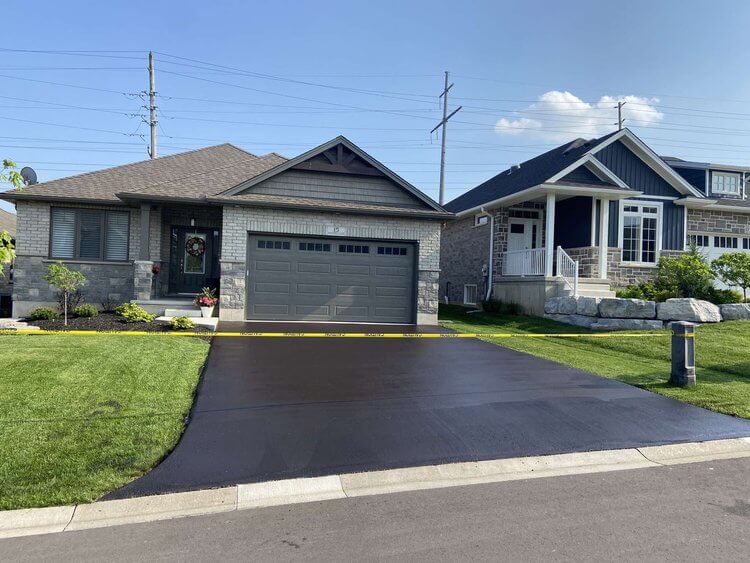 A newly-paved asphalt driveway in front of a single-story house, marked off with yellow caution tape. The house has a grayish exterior with a garage door and a well-maintained lawn. Another similar house is adjacent to it, separated by a small grassy area.