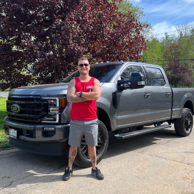 A person with sunglasses, arm tattoos, and a red sleeveless shirt is standing with arms crossed in front of a large gray Ford truck on an asphalt driveway. The driveway is bordered by grass and trees with dark red and green leaves. The sky is clear and sunny.