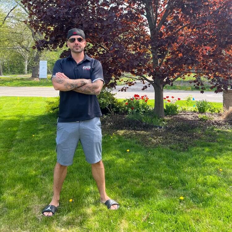 A man in sunglasses stands on a grassy lawn with his arms crossed. He is wearing a black polo shirt, gray shorts, black sandals, and a backwards cap. Behind him is an asphalt path leading to a garden bed with red flowers and a tree with dark red leaves.