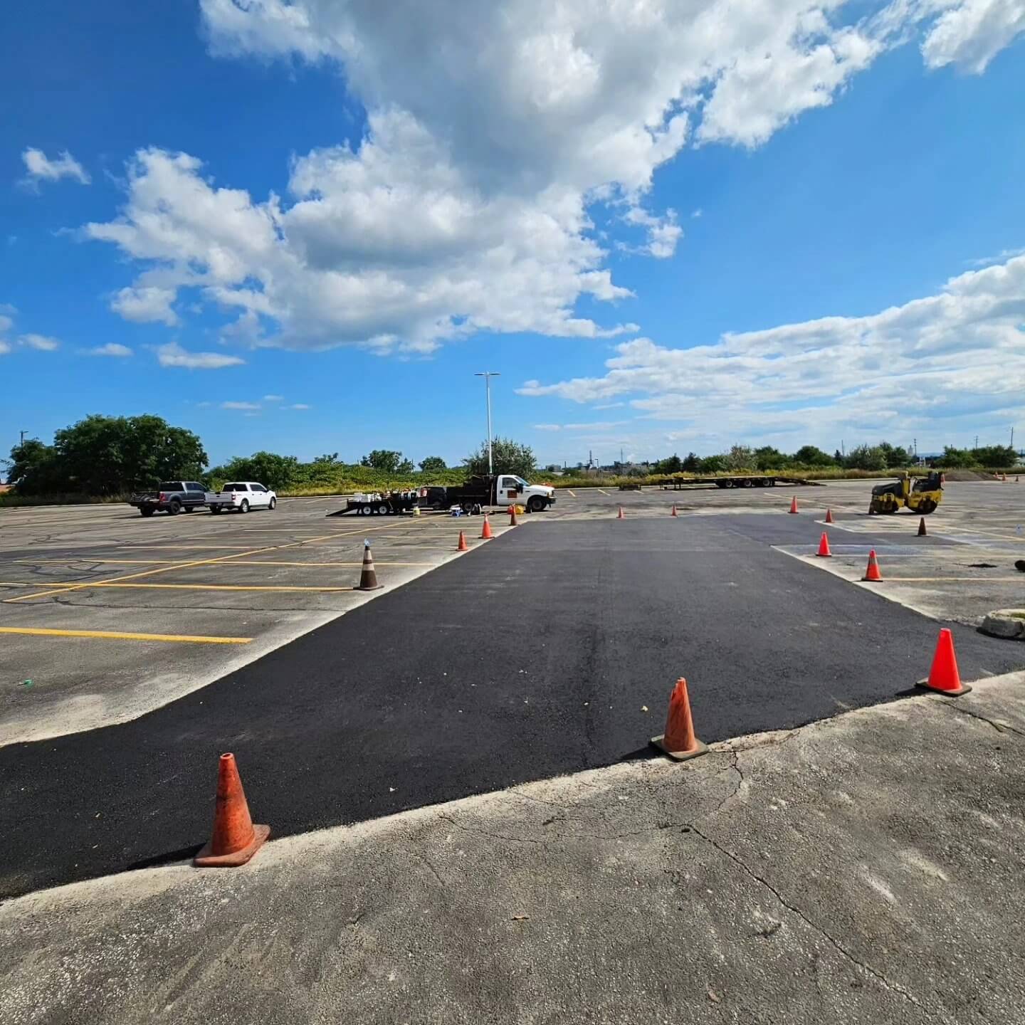 A parking lot under construction, featuring fresh paving with a newly laid black asphalt section bordered by orange traffic cones. Several vehicles are parked in the background, and a blue sky with scattered clouds is overhead.