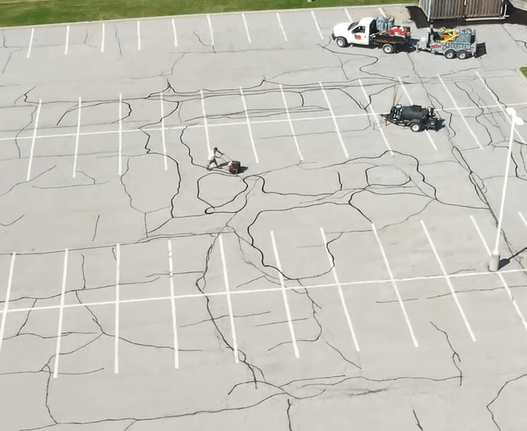 A large, empty parking lot with numerous visible cracks on the asphalt. Two vehicles are parked on the far side with equipment, and one person is working on repairing cracks. Several parking spaces are marked with white lines, much like a worn driveway in need of maintenance.