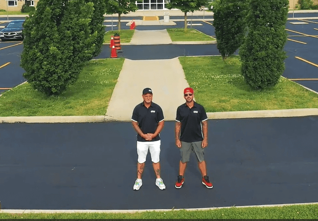 Two men stand smiling in a mostly empty parking lot on a sunny day, the fresh asphalt glinting beneath their feet. Both are wearing black polo shirts and caps. One man has on white shorts and sneakers, while the other wears gray shorts and black sneakers. Green trees and grass are visible in the background.