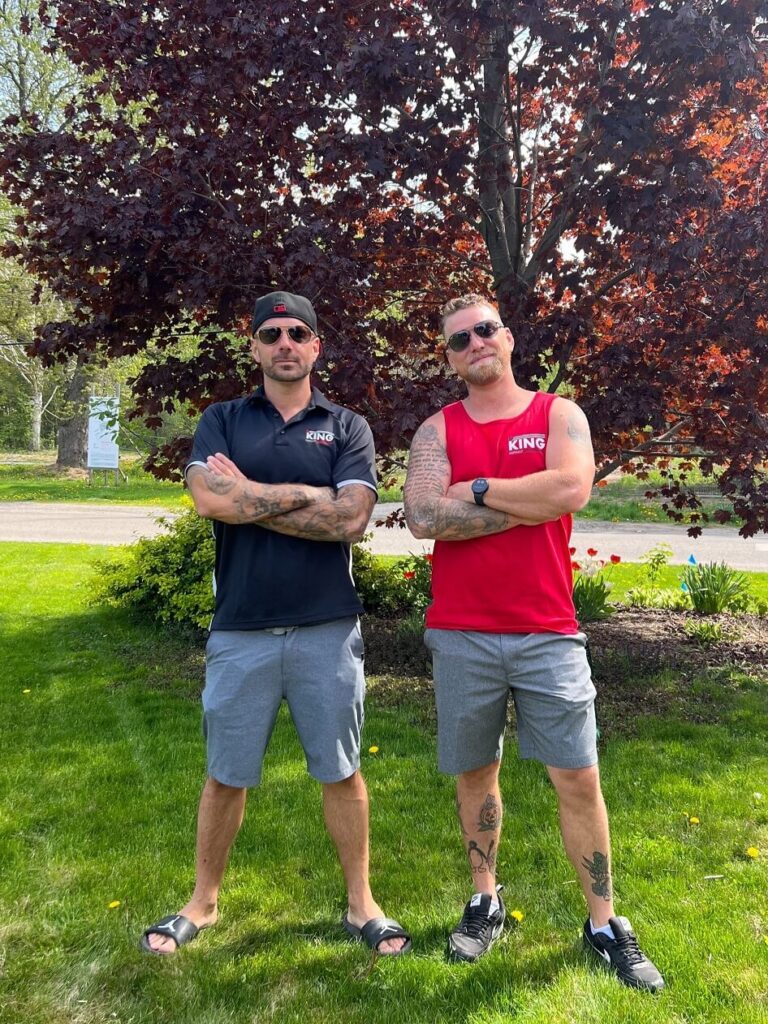 Two men stand on grass with arms crossed in front of a tree with reddish leaves. On the left, a man wears a black hat, sunglasses, a black shirt, and gray shorts. His companion sports sunglasses, a red sleeveless shirt, and gray shorts. Both men have tattoos and look ready for paving the asphalt nearby.