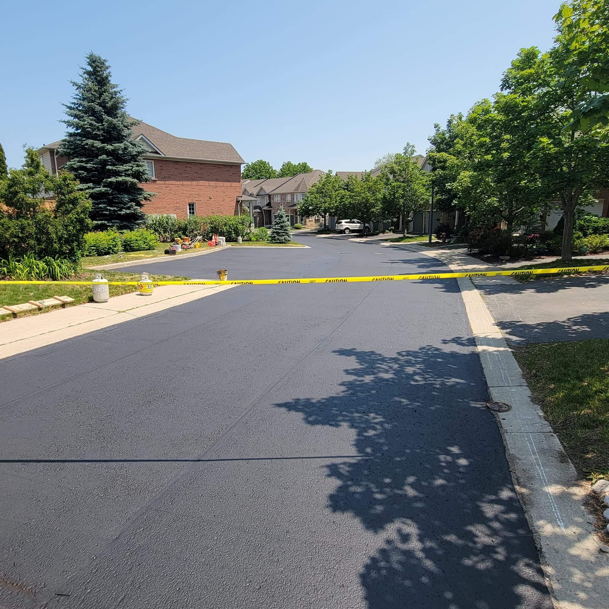 A commercial lot in Ancaster with fresh asphalt is blocked off with yellow caution tape. Brick houses, each featuring a tidy driveway, and green trees line the street under a clear blue sky. The scene, marked by recent paving work, is peaceful with no cars or people visible.