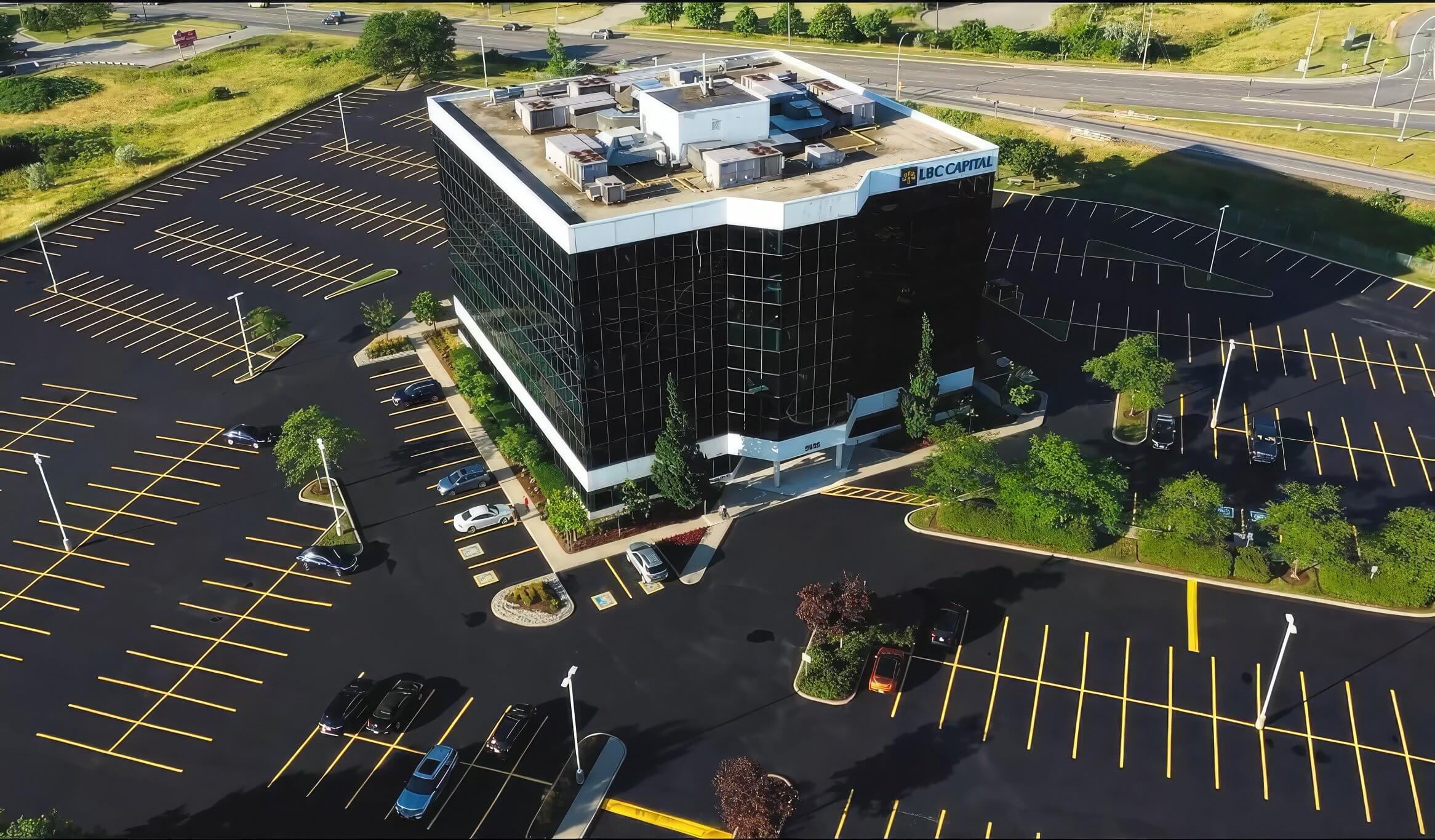 Aerial view of a multi-story office building in Burlington surrounded by an expansive parking lot with numerous marked parking spaces. The asphalt paving accentuates the reflective glass facade, trees, and landscaped greenery. Several cars are parked near the building, and a highway is visible in the background.