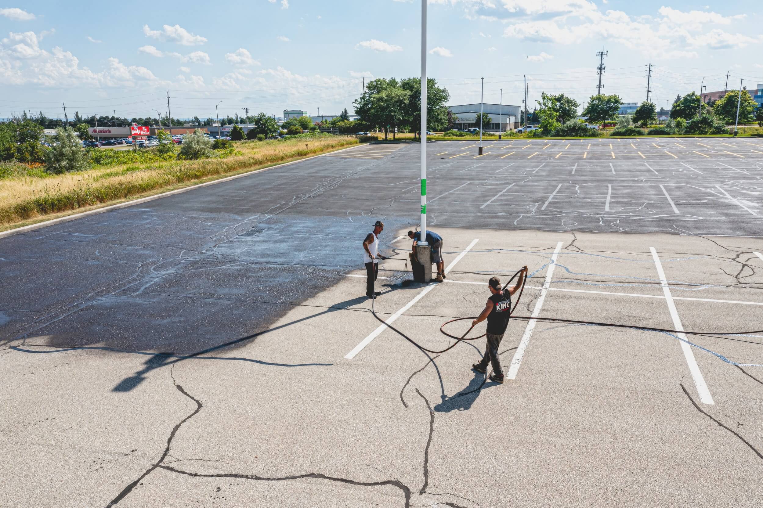 Two workers are seen power washing a large, empty parking lot. One worker operates the pressure washer while the other manages the hose. The partially wet and cracked parking lot, with patches of greenery in the background under a clear blue sky, looks ready for paving or driveway maintenance.