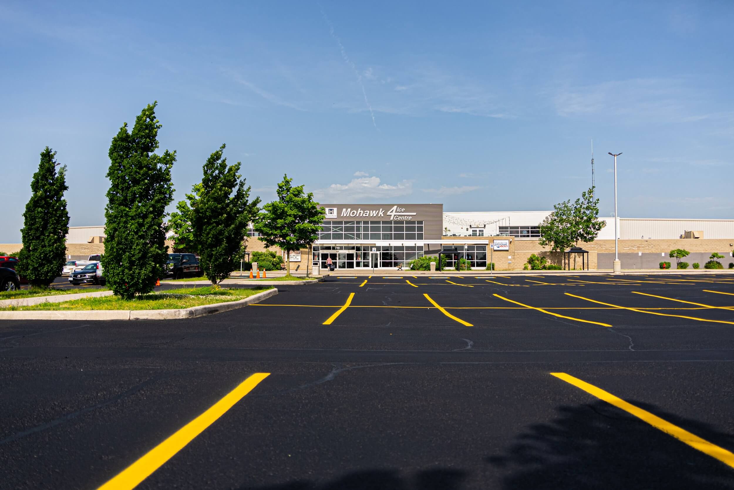 A newly paved, empty parking lot stretches out in front of a large industrial building with a "Mohawk" sign on it. Young trees line the left side of the parking lot, and the sky above is clear and blue with a few wispy clouds.