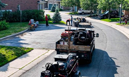 A residential neighborhood street is undergoing maintenance work. Several workers wearing safety vests are gathered around a trailer loaded with paving equipment. Another trailer with asphalt machinery is parked ahead, and houses with trees line both sides of the street.