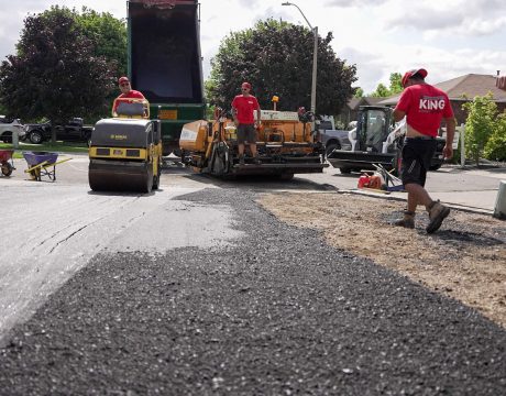 Workers in red shirts and caps repave a residential street. A dump truck pours asphalt into a paver, while a worker operates a steamroller to smooth the surface. Trees and houses line the background as the paving progresses smoothly.