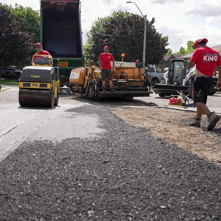 Workers in red shirts are paving a residential road in Hamilton, Ontario. A steam roller is compacting the fresh asphalt, while an asphalt paver machine and a dump truck are in the background. Equipment and tools are scattered around, with houses and trees lining the street. It's similar to laying a new driveway or parking lot transition.