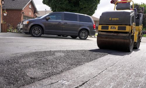 A road roller compacts freshly laid asphalt on a residential street, ensuring smooth paving. A parked minivan and brick houses with lawns and flower baskets are visible in the background.