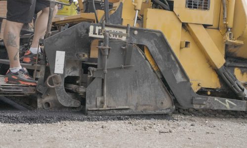 A close-up of a yellow paving machine in action. The lower part of the asphalt machine is laying down a new layer of black pavement. A person wearing black shorts and red and black sneakers is standing on the machine, controlling it.