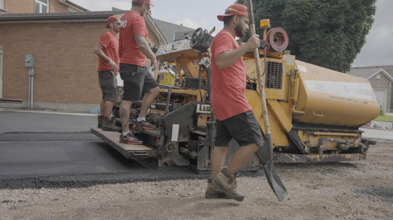 Three asphalt workers in red shirts and caps are paving a road using heavy machinery. One worker stands on the machine, another operates it, and a third rakes the fresh asphalt. A house and trees are visible in the background.