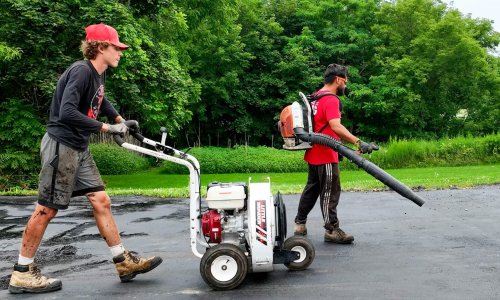 Two workers are seen asphalt sealing a driveway. One worker, on the left, is pushing a seal coating machine, while the other, on the right, is operating a blower backpack. Both are wearing casual work clothes and are surrounded by green trees in the background, reminiscent of a parking lot being paved.