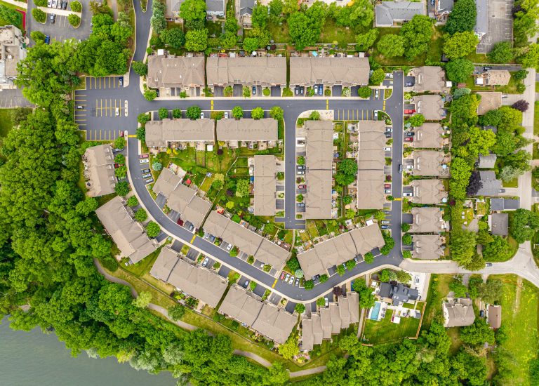 Aerial view of a residential neighborhood with rows of houses surrounded by green trees. The houses are organized in a grid pattern with clear streets and asphalt driveways. At the bottom left corner is a body of water partially visible, bordered by more trees.