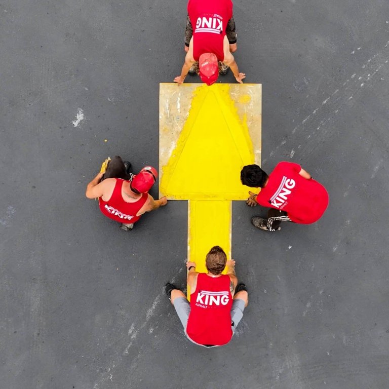 Four people in red shirts marked "KING" work together to paint a large yellow arrow on the gray asphalt. The photo, taken from above, shows the team positioning a wooden board to create the arrow shape in what appears to be a parking lot.