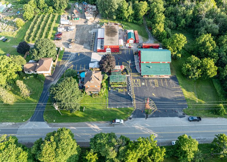 Aerial view of a property with several buildings surrounded by trees. The area includes a parking lot with clearly marked asphalt spaces, a large warehouse, smaller buildings, and vehicles. There's a road in the foreground and various green spaces and landscaping.