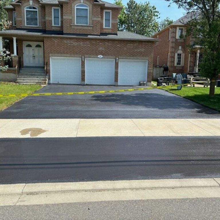 A brick two-story house with three white garage doors has a freshly paved asphalt driveway. Yellow caution tape is placed across the driveway, indicating that it is not yet ready for use. Nearby trees and neighboring houses are visible.