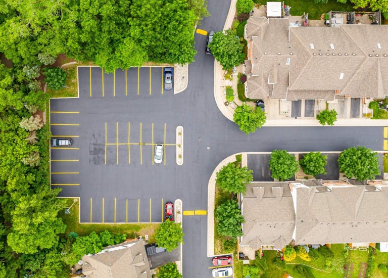 Aerial view of a nearly empty residential parking lot with a few parked cars on smooth asphalt. The surroundings are lush with greenery. Adjacent to the parking lot are residential buildings, sidewalks, and well-maintained landscaping.