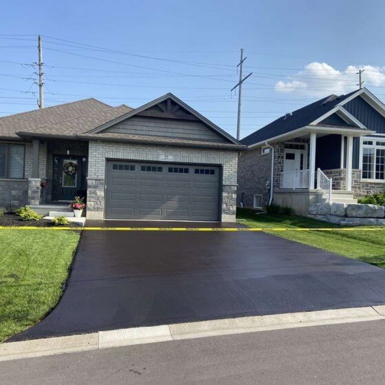 A newly-paved asphalt driveway in front of a single-story house, marked off with yellow caution tape. The house has a grayish exterior with a garage door and a well-maintained lawn. Another similar house is adjacent to it, separated by a small grassy area.