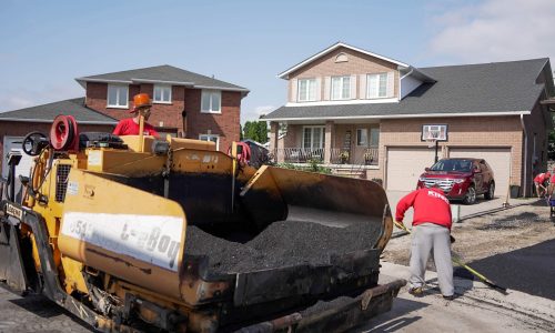 Construction workers in red shirts operate machinery to pave a residential street with asphalt in front of brick houses. One worker is on the machine while the other meticulously spreads paving material on the ground. A red SUV is parked in a driveway in the background.