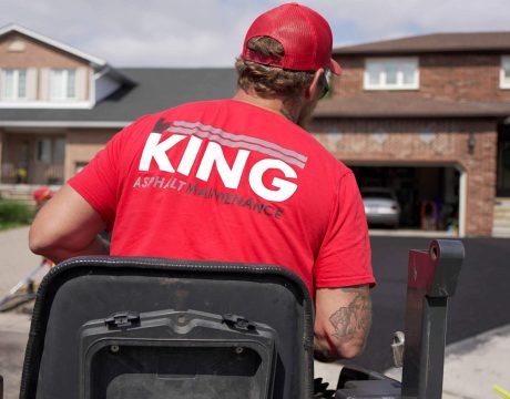 A worker in a red "KING Asphalt Maintenance" shirt and cap operates an asphalt paving machine while working on a driveway in front of a two-story brick house with a double garage. The scene is set in a suburban neighborhood on a sunny day.