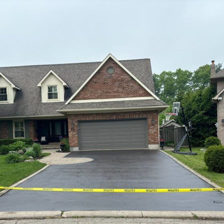 A suburban house with a beige and red brick exterior, two stories, and a neatly paved driveway. Yellow caution tape blocks entry to the driveway, which features a basketball hoop on the side. Green bushes and trees surround the property, emphasizing its well-maintained appearance.