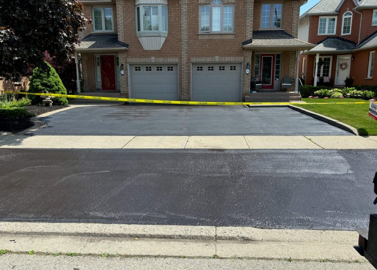 A freshly paved driveway in front of a brick townhouse is cordoned off with yellow caution tape. The smooth, shiny black asphalt from the new paving extends across the two garages with white doors. Green lawns and neighboring houses are visible, giving a pristine look to the parking lot area.