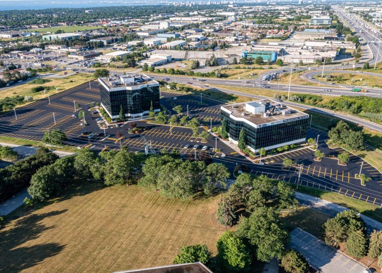 Aerial view of two office buildings with glass exteriors in a business park, surrounded by large paved parking lots and green spaces. A highway encircles the area, and a cityscape is visible in the background. Sparse cars are parked on the asphalt lots.