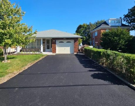 A single-story brick house with a freshly paved asphalt driveway leading to a white garage door. There's a small garden area to the left with a tree and shrubs, and another house is visible to the right. The sky is clear and blue.