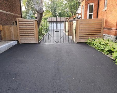 A freshly paved driveway leads to a wooden and metal gate between two brick houses. Behind the gate, there's a white garage surrounded by trees and greenery. The gate, with vertical wooden slats and a minimalist metal design in the center, complements the smooth asphalt underfoot.