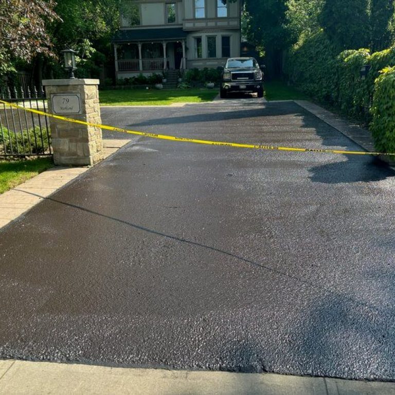 A freshly paved driveway is blocked off with yellow caution tape. The driveway leads to a residence with a black vehicle parked near the garage. The surroundings include green trees and bushes, and the entrance has stone pillars, showcasing meticulous paving work.