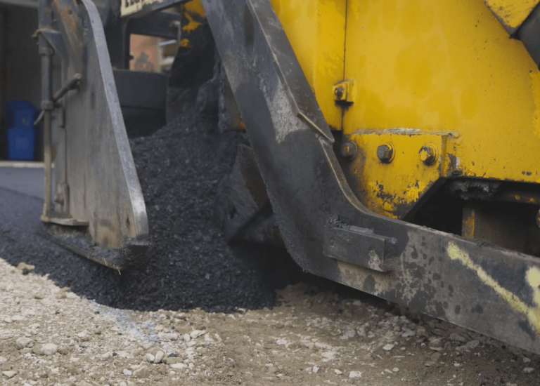 A driveway paving machine lays fresh asphalt at a Hamilton residence. The yellow machinery is partially visible, with black asphalt being poured smoothly onto the ground, paving the way for a pristine surface. A car and a garage are seen in the background.