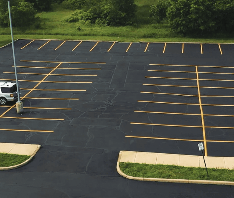 Aerial view of an almost empty parking lot with freshly paved asphalt, featuring a single white SUV parked near the left side and a dark blue car on the right. The lot has newly painted yellow lines and is surrounded by green grass and trees.