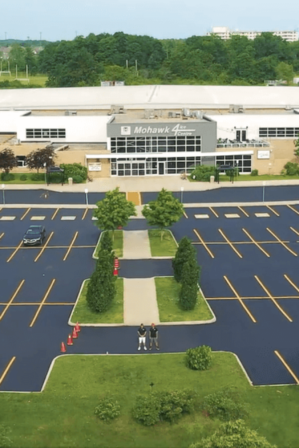 A large building with a sign reading "Mohegan Arena" is surrounded by a mostly empty parking lot paved with asphalt. Only a few parked cars are visible along the driveway. The area is landscaped with trees and bushes, and there is a walkway leading to the entrance. The background shows a suburban area with greenery.