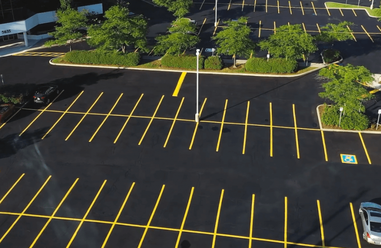 Aerial view of a nearly empty asphalt parking lot with clearly marked yellow lines for parking spaces, including designated handicapped spots. There are a few scattered cars, green trees, and a building visible at the edge of the paved lot.