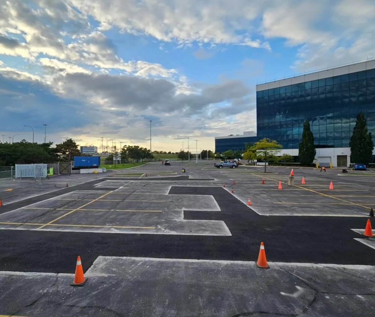 A parking lot with orange traffic cones arranged in a pattern, possibly for a driving course, sits adjacent to a large modern glass building under partly cloudy skies. A few cars are scattered around and trees frame the background, all set against the smooth expanse of asphalt.