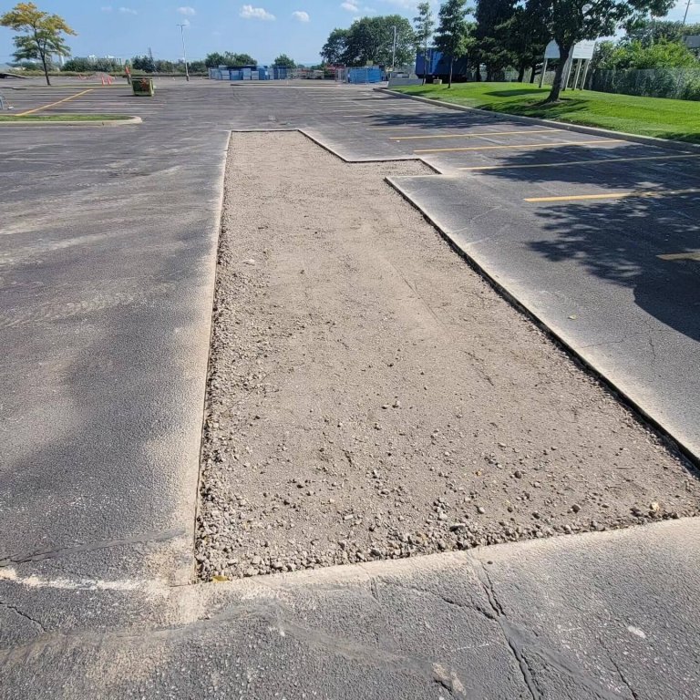 A section of an asphalt parking lot has been cut out, exposing a rectangular area of dirt and gravel underneath. The surrounding lot is empty and painted with parking lines. Trees and a grassy area are visible in the background under a clear blue sky, awaiting new paving.