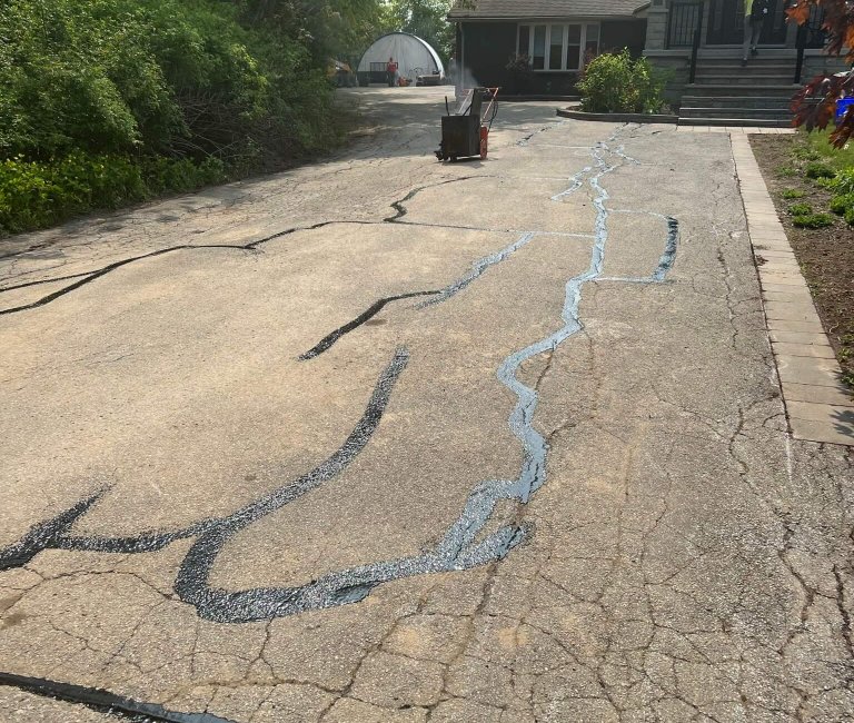 A paved driveway with visible cracks filled with fresh sealant stretches toward a house with a small staircase and a covered porch. A wheelbarrow brimming with paving tools sits in the middle, while trees and bushes line one side, lending a serene touch to the scene.