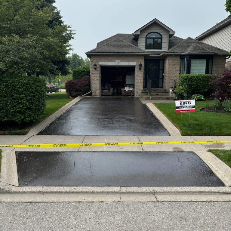 A house with a freshly paved driveway in Ancaster Ontario, cordoned off with yellow caution tape. An "KING" contractor sign stands on the right side of the asphalt driveway. The garage door is open, partially revealing the interior. Shrubs and trees surround the property.