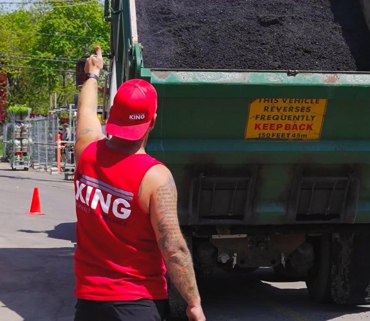 A person in a red hat and red shirt with "KING" printed on the back is directing a truck that's dumping a load of asphalt. The truck has a sign that reads, "This vehicle reverses frequently. Keep back 150 feet / 45m." Nearby, safety cones and a fence ensure the paving site remains secure.