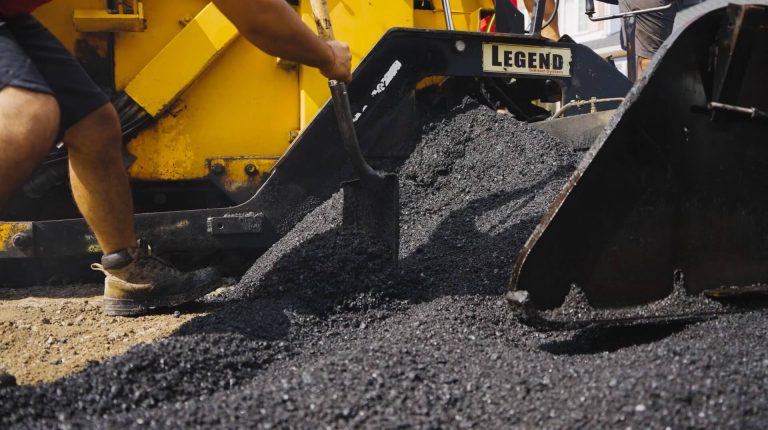 A worker operates heavy machinery labeled "Legend," spreading hot asphalt during a road construction project. The yellow machine is involved in precise paving as two large shovels move the asphalt onto the ground.