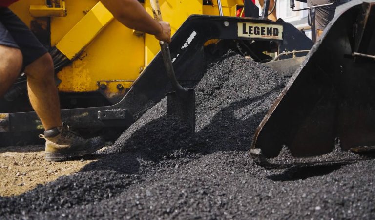 A worker uses a tool to spread fresh asphalt being laid by yellow paving machinery, likely for a new parking lot. The black and granular asphalt is labeled "LEGEND." Wearing boots, the worker's arm and part of their torso are visible as they diligently work.