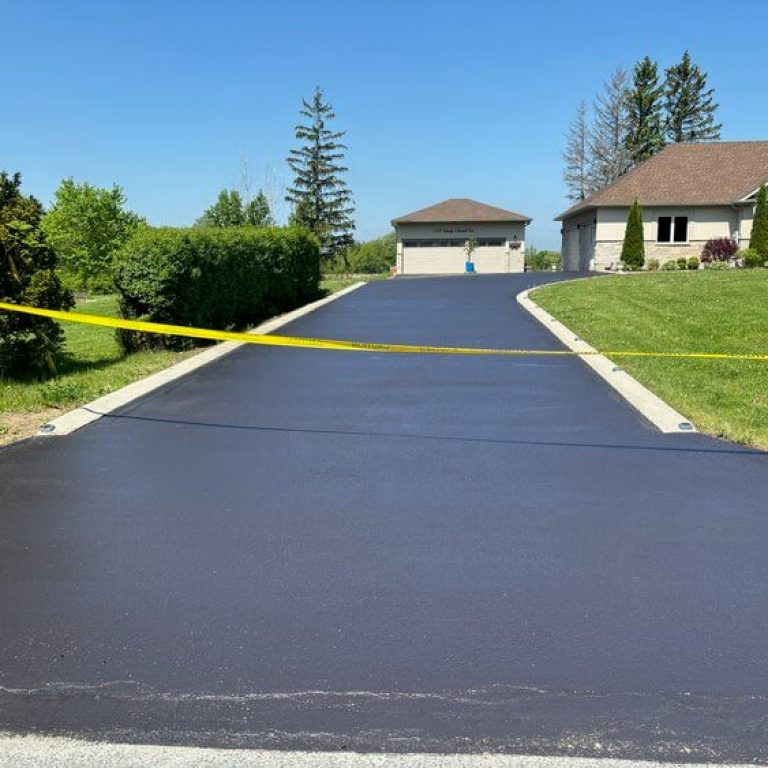 A newly paved asphalt driveway leading to a house, roped off with yellow caution tape. The driveway is bordered by green grass and landscaping. In the background, a garage and house are visible under a clear blue sky.