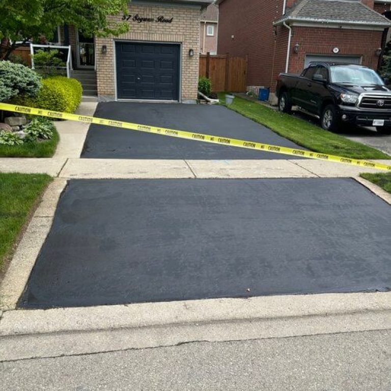 A freshly paved asphalt driveway in front of a house is blocked off with yellow caution tape. To the right, a black truck is parked in the new driveway. The house, featuring brick siding and a double garage door, stands behind this neat scene.