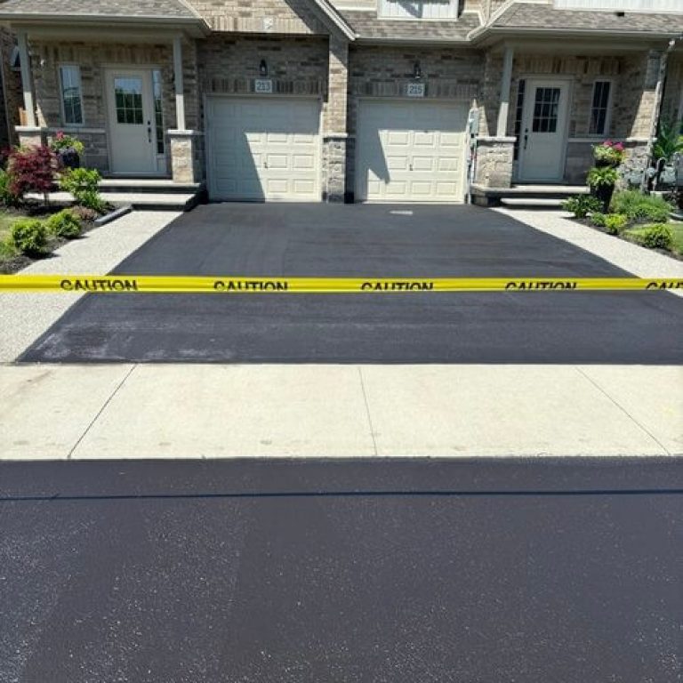 A newly paved asphalt driveway in front of a two-story brick house is blocked by caution tape. The two garages have identical doors, and there are small landscaped garden areas with plants and shrubs on either side of the driveways.