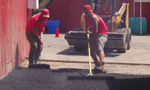 Two construction workers wearing red shirts and caps are spreading asphalt with rakes, diligently paving near a red building. Behind them, another worker operates a yellow skid-steer loader. Safety cones and various equipment are visible in the background under a sunny sky.