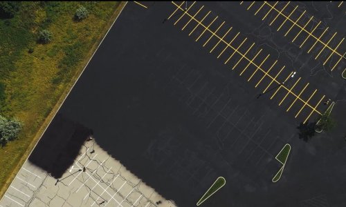 Aerial view of a large, mostly empty parking lot. The asphalt appears freshly paved except for one worn-out section in the bottom left corner with visible cracks. Bright yellow parking lines are neatly painted across the surface, and a grassy area borders one side.