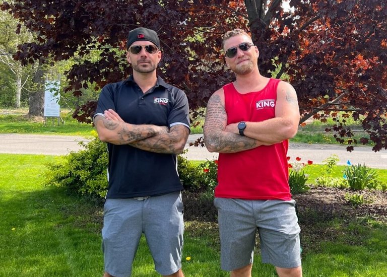 Two men stand on grass with arms crossed in front of a tree with reddish leaves. On the left, a man wears a black hat, sunglasses, a black shirt, and gray shorts. His companion sports sunglasses, a red sleeveless shirt, and gray shorts. Both men have tattoos and look ready for paving the asphalt nearby.