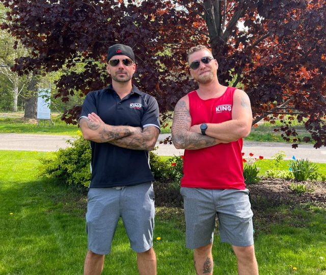 Two individuals standing on grass in front of a tree with dark red leaves. Both have tattoos and wear sunglasses, shorts, and sandals. The person on the left wears a black shirt and cap, while the person on the right wears a red sleeveless shirt, near an area where asphalt meets concrete paving.